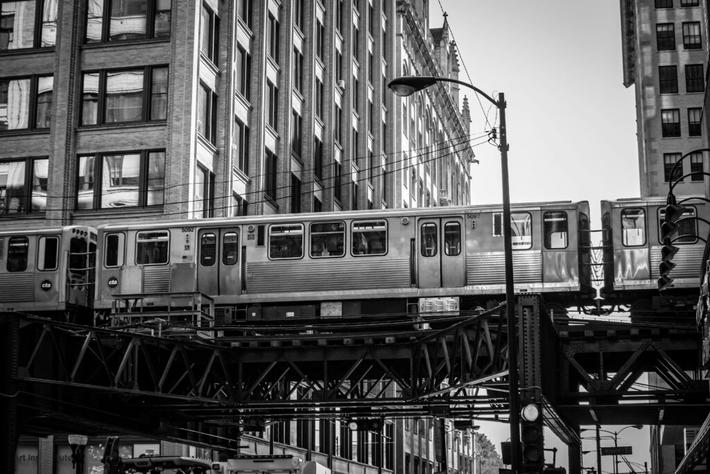 A Chicago L train travels past historic buildings in a black and white cityscape.